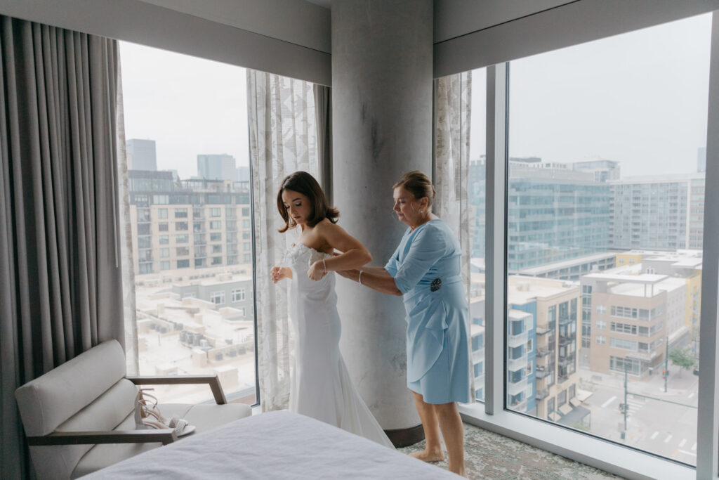 bride's mother helping bride strap wedding dress on standing in front of glass window overlooking city