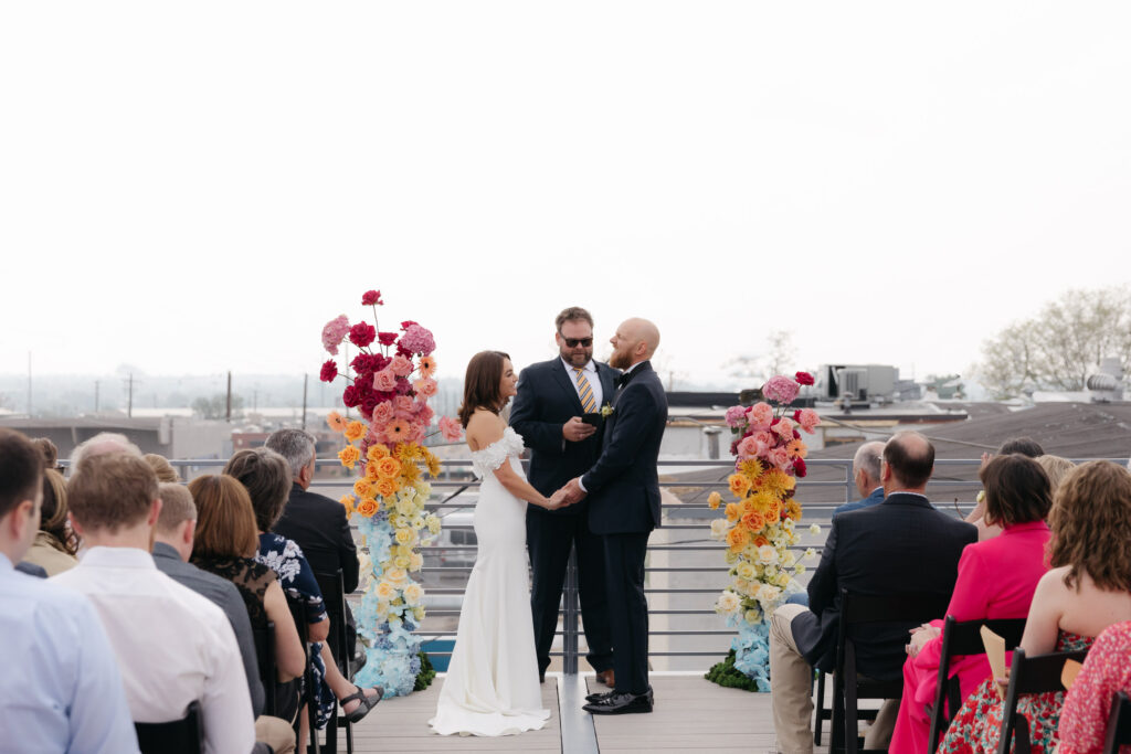 bride and groom laughing during ceremony and standing in between bright, colorful floral fixture