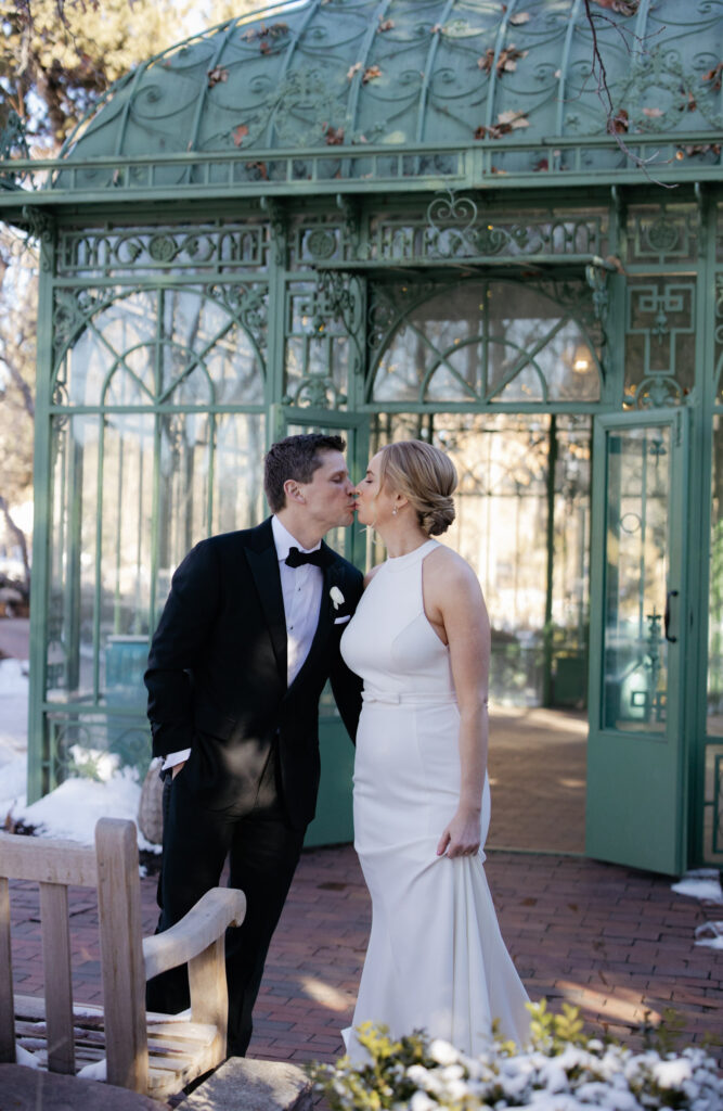 Groom kisses bride while standing in front of Woodlands Mosaic Solarium during Denver wedding photos