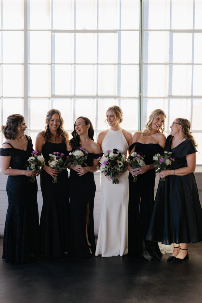 Bridesmaids and bride candidly laughing in front of large, industrial windows during Denver wedding photos at Moss venue