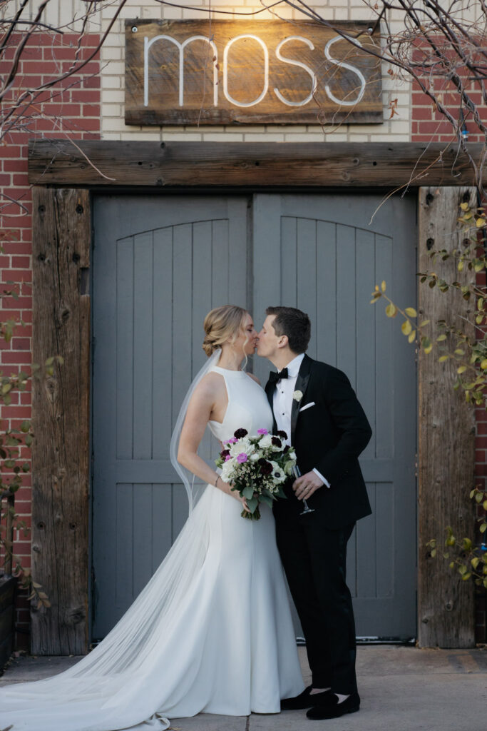 Bride and groom kissing in front of industrial, wood doors of Denver wedding venue Moss