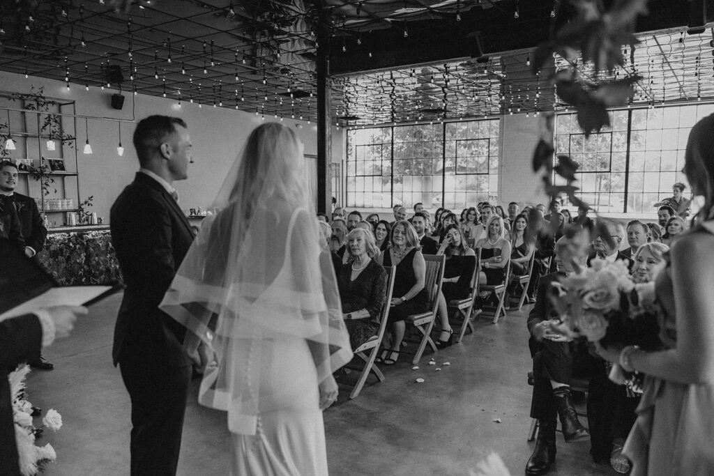 bride and groom facing audience of guests during wedding ceremony inside industrial venue