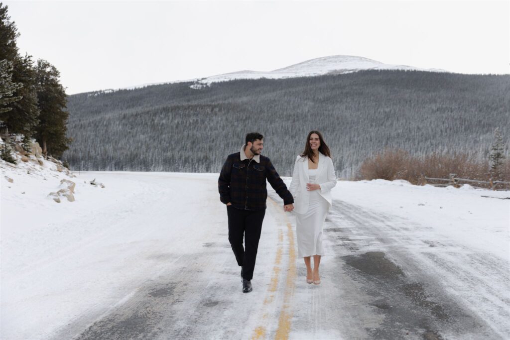 Couple holding hands walking down middle of snow filled road during Colorado engagement photos at Echo Lake by Colorado engagement photographer Caroline Brackney