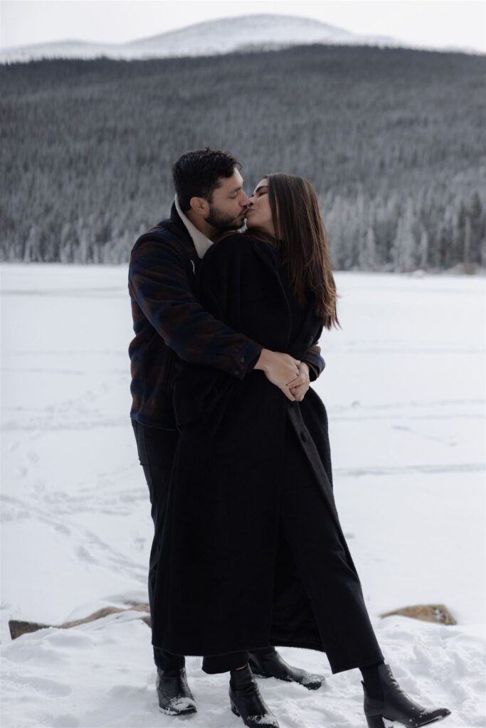 Couple embrace in kiss standing in front of frozen lake and snowy forest
