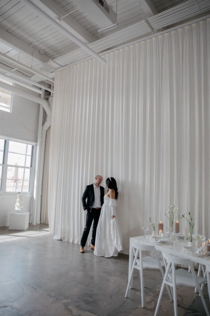 couple standing in front of tall white backdrop in natural light studio in wedding attire