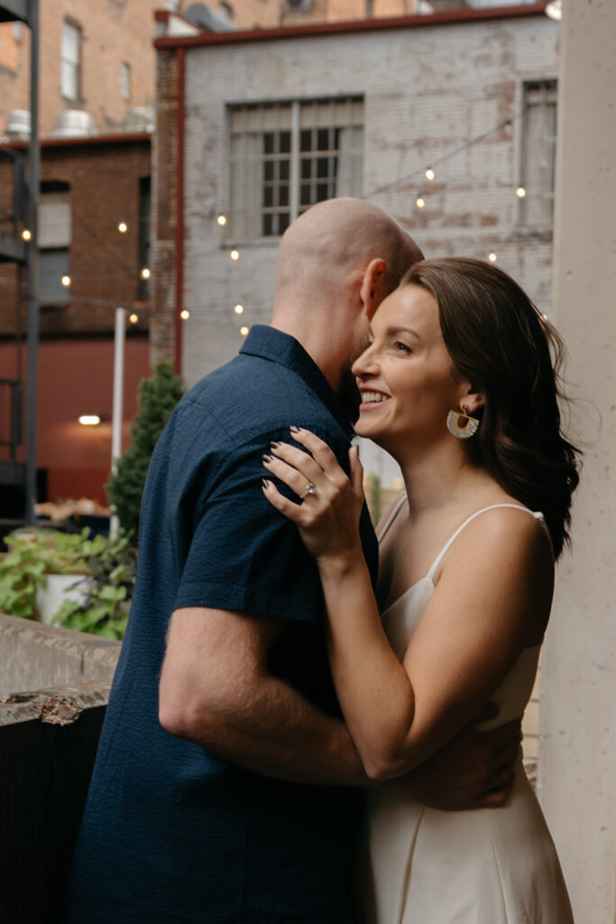 couple standing in front of twinkle lights and brick buildings while taking engagement photos