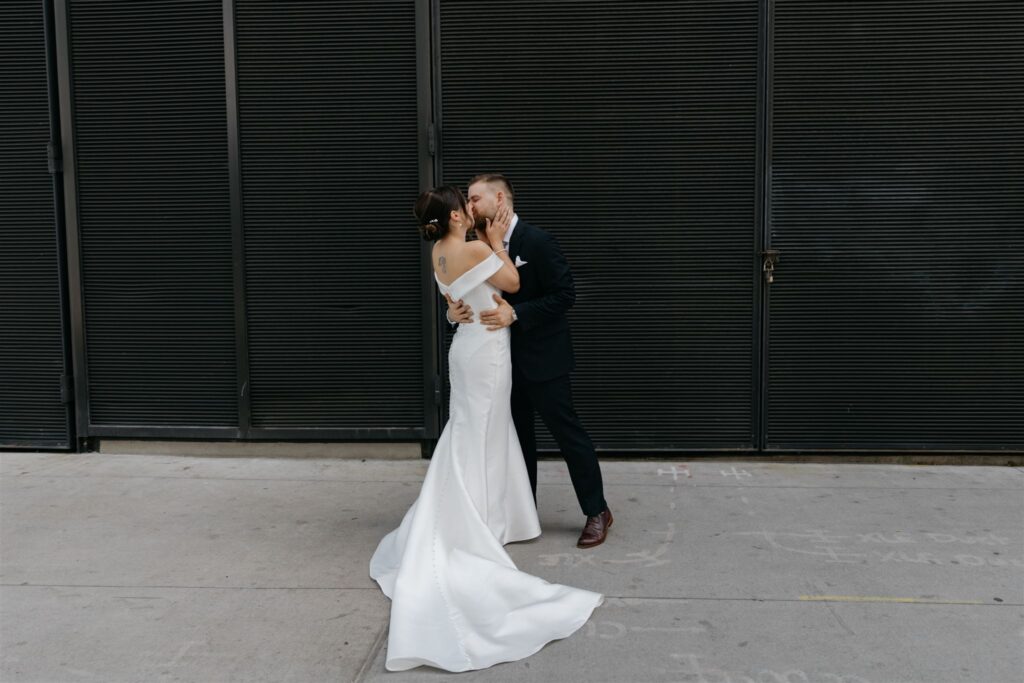Bride and groom embracing in a kiss standing on sidewalk in front of dark industrial, wall
