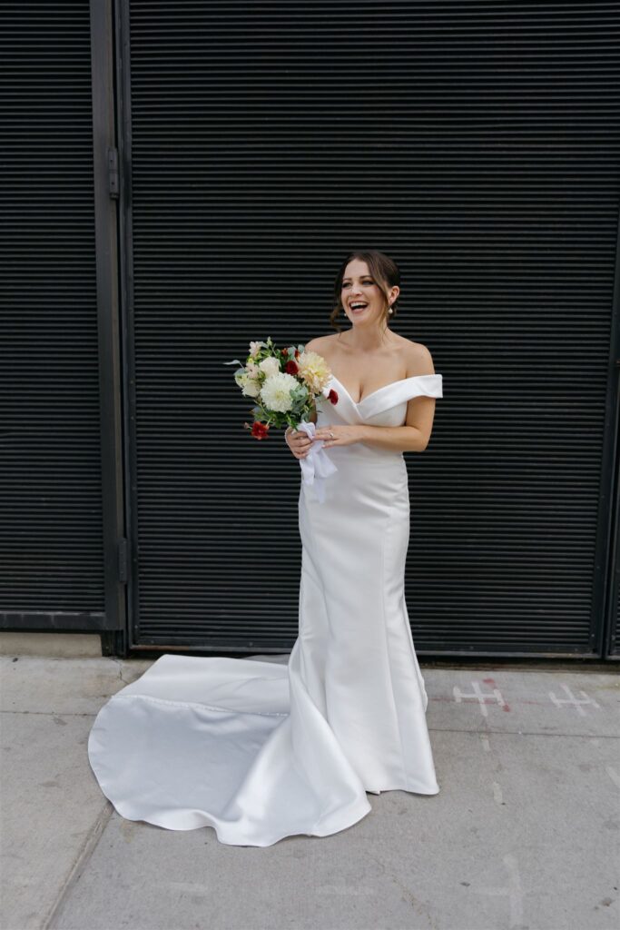 bride in off shoulder wedding dress holding bouquet and standing on concrete sidewalk with industrial wall behind her