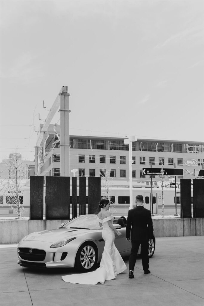 black and white image of bride and groom walking towards convertible sports car with trains and Denver Union Station in background
