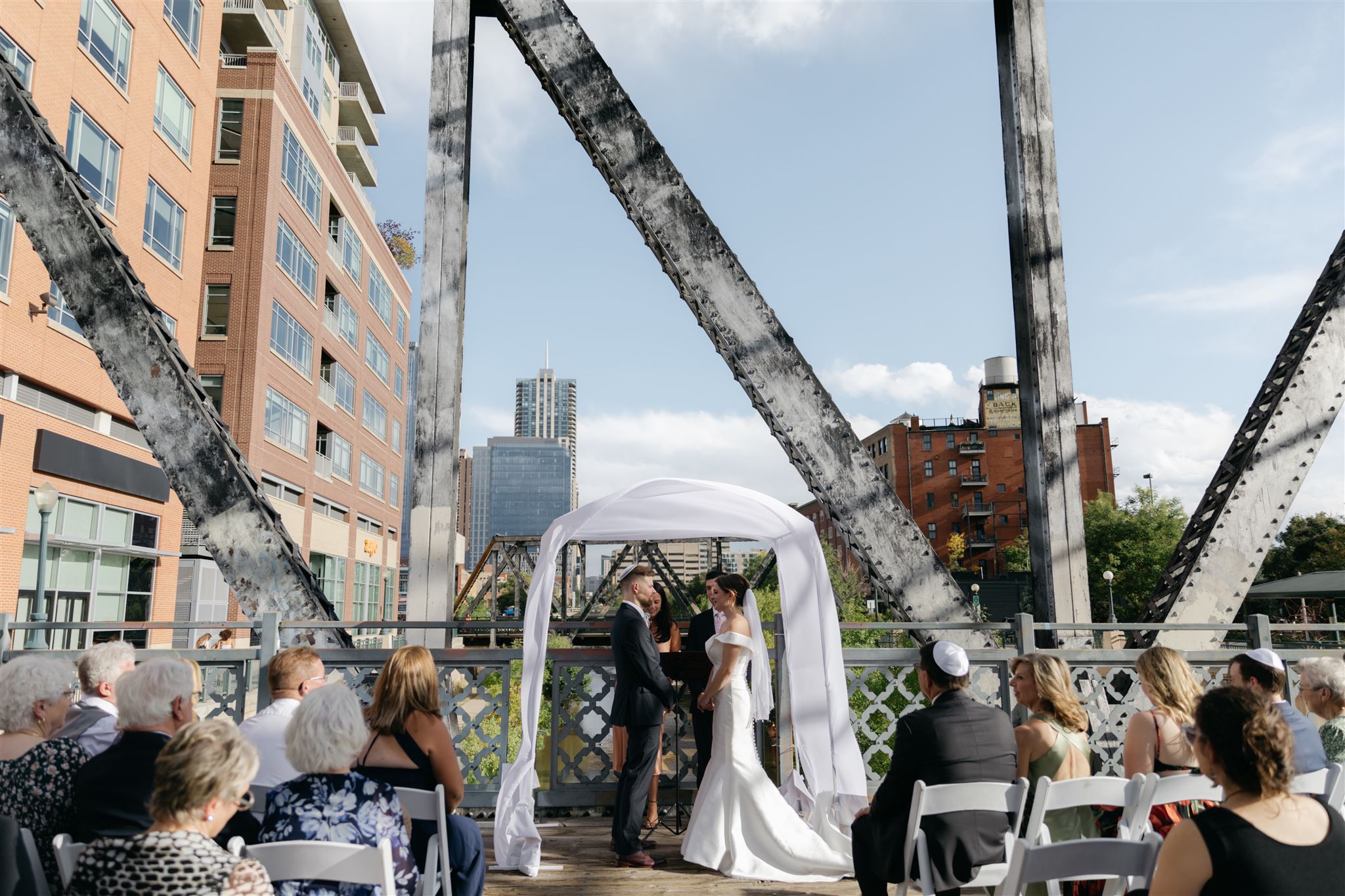 A bride and groom standing under a chuppah on Wewatta bridge at Coohills restaurant in downtown Denver with tall buildings in background