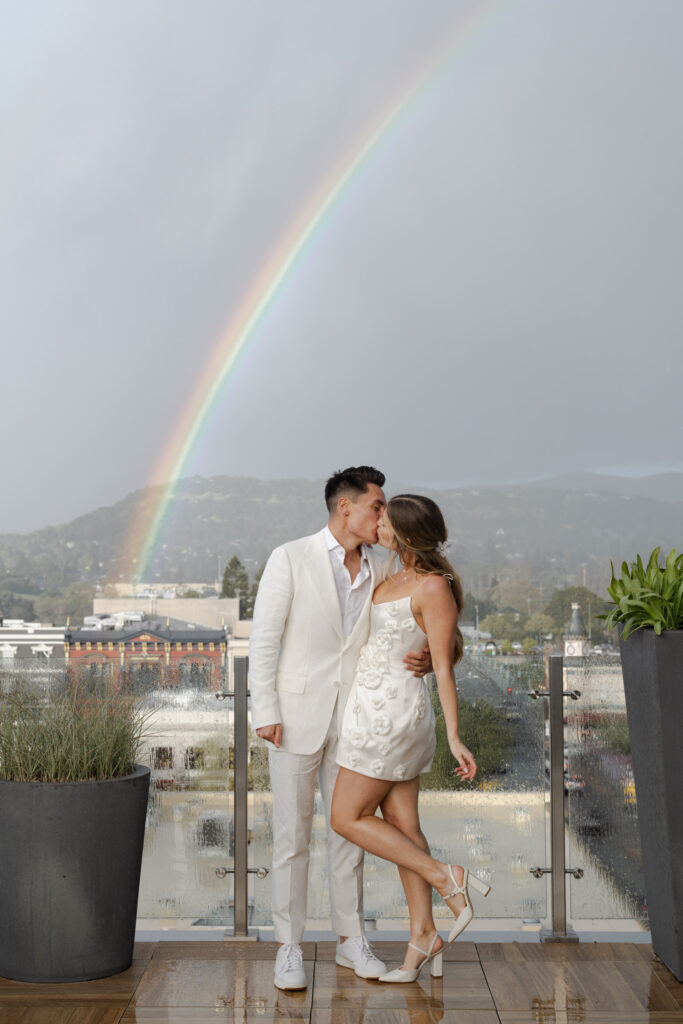 Rainbox backdrops sky while couple kisses on rooftop of wedding welcome party on rooftop