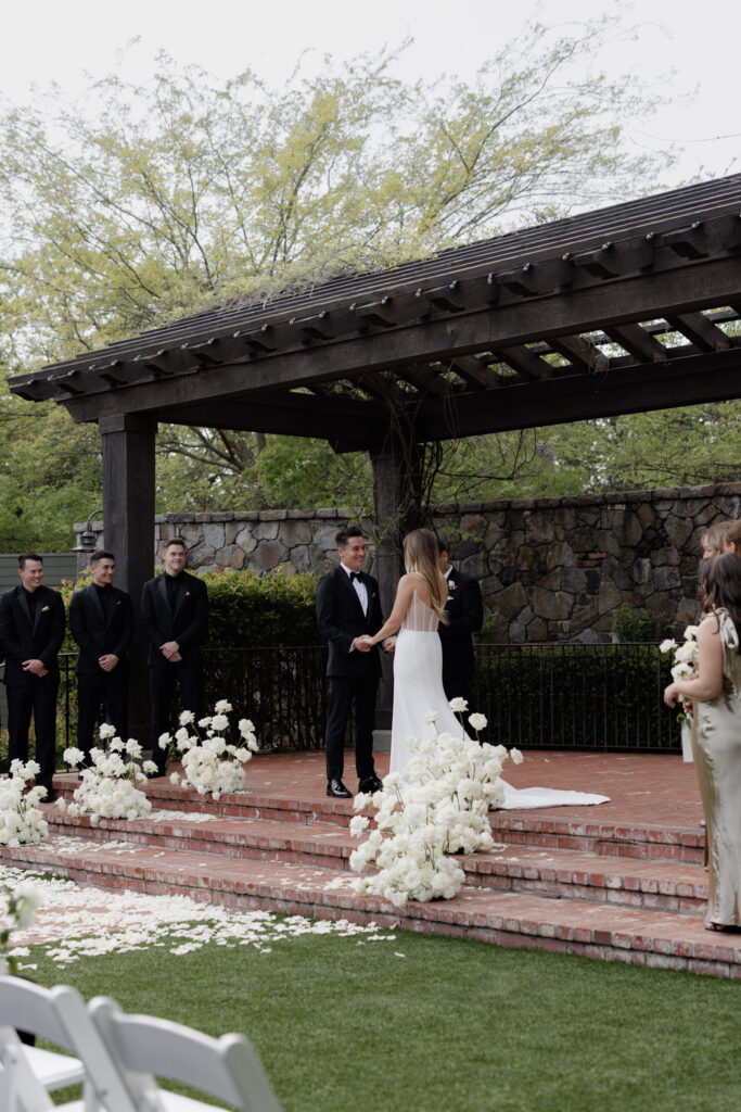 Bride and groom stand between white roses during courtyard wedding ceremony at the Estate Yountville